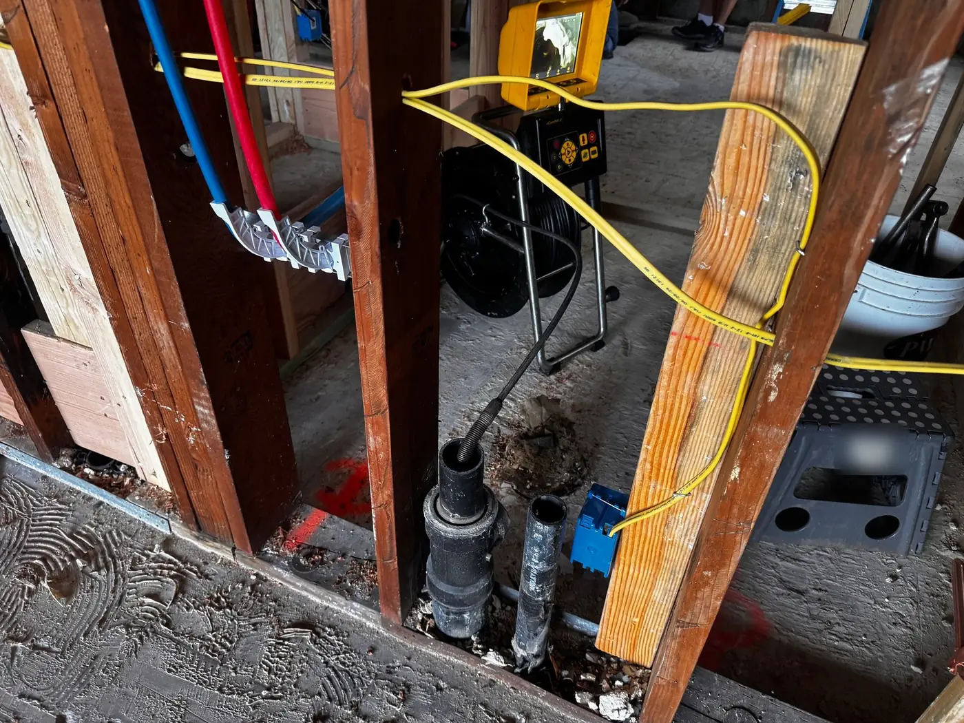 Sewer camera push cable entering an open drain pipe at floor level inside a house under remodel, with the camera reel and monitor behind the wall framing