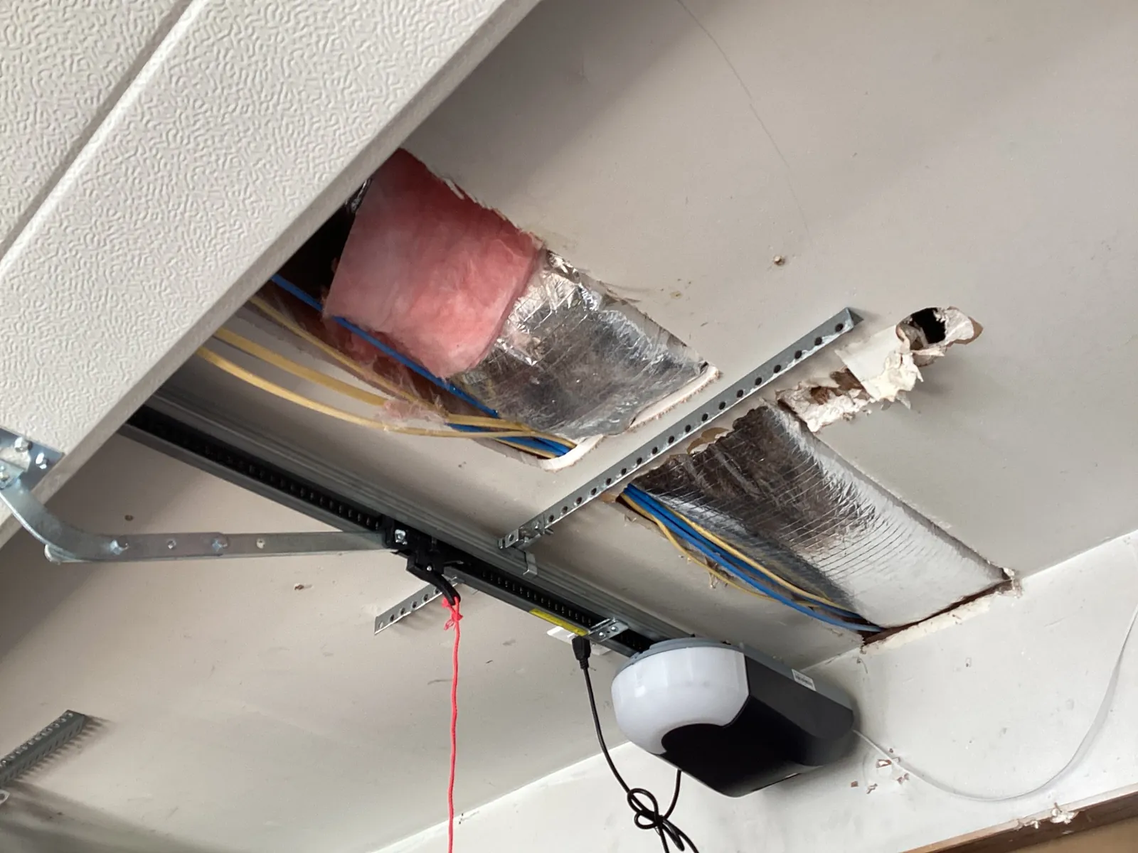 Garage ceiling with two large rectangular openings cut in the drywall, exposing foil-wrapped flexible ducts, pink insulation and wiring, plus a ragged hole beside a garage door opener bracket