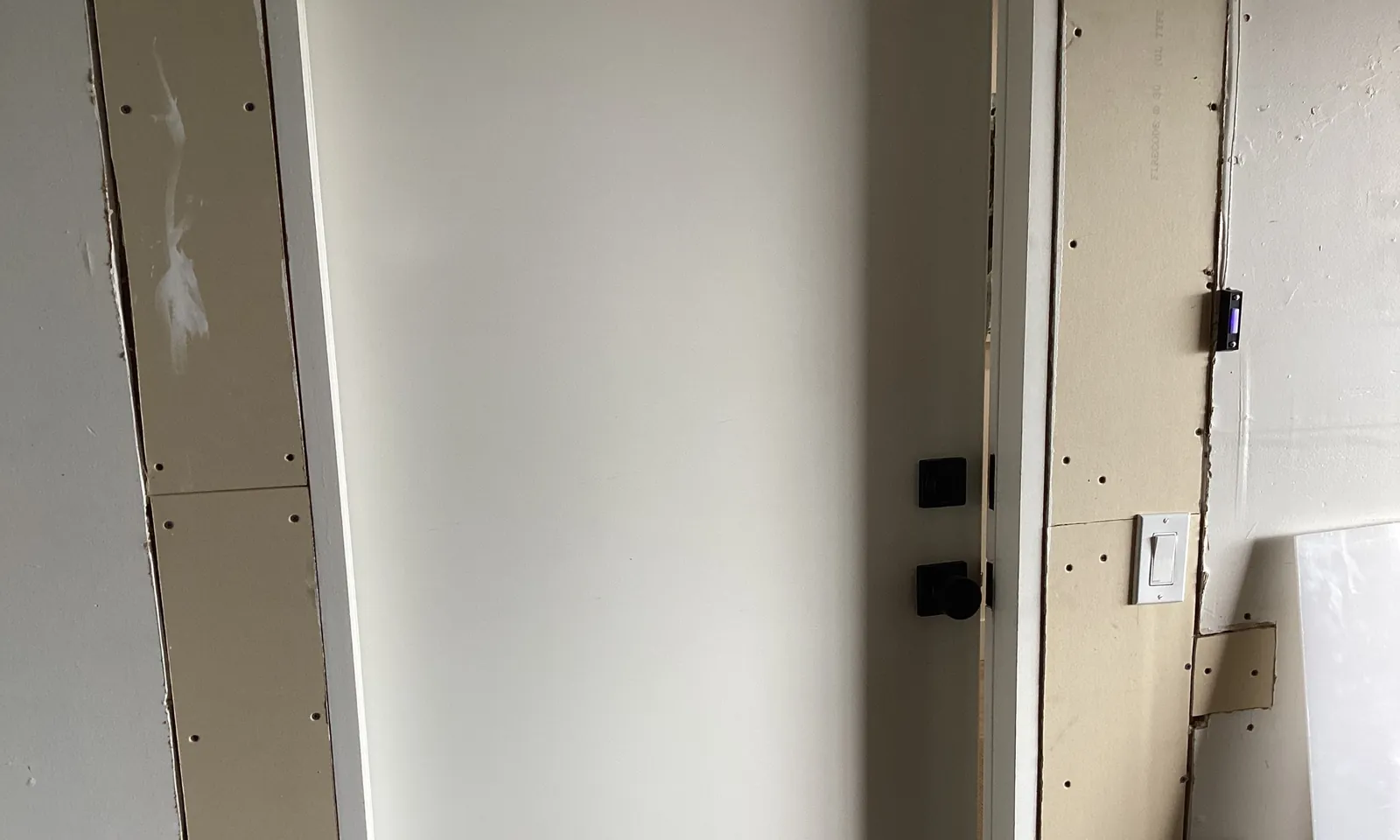 Door from the garage into the house, standing slightly open, with fresh drywall strips around the frame stamped Firecode Type X, screw heads and seams left unfinished, and a separate small patch beside the light switch