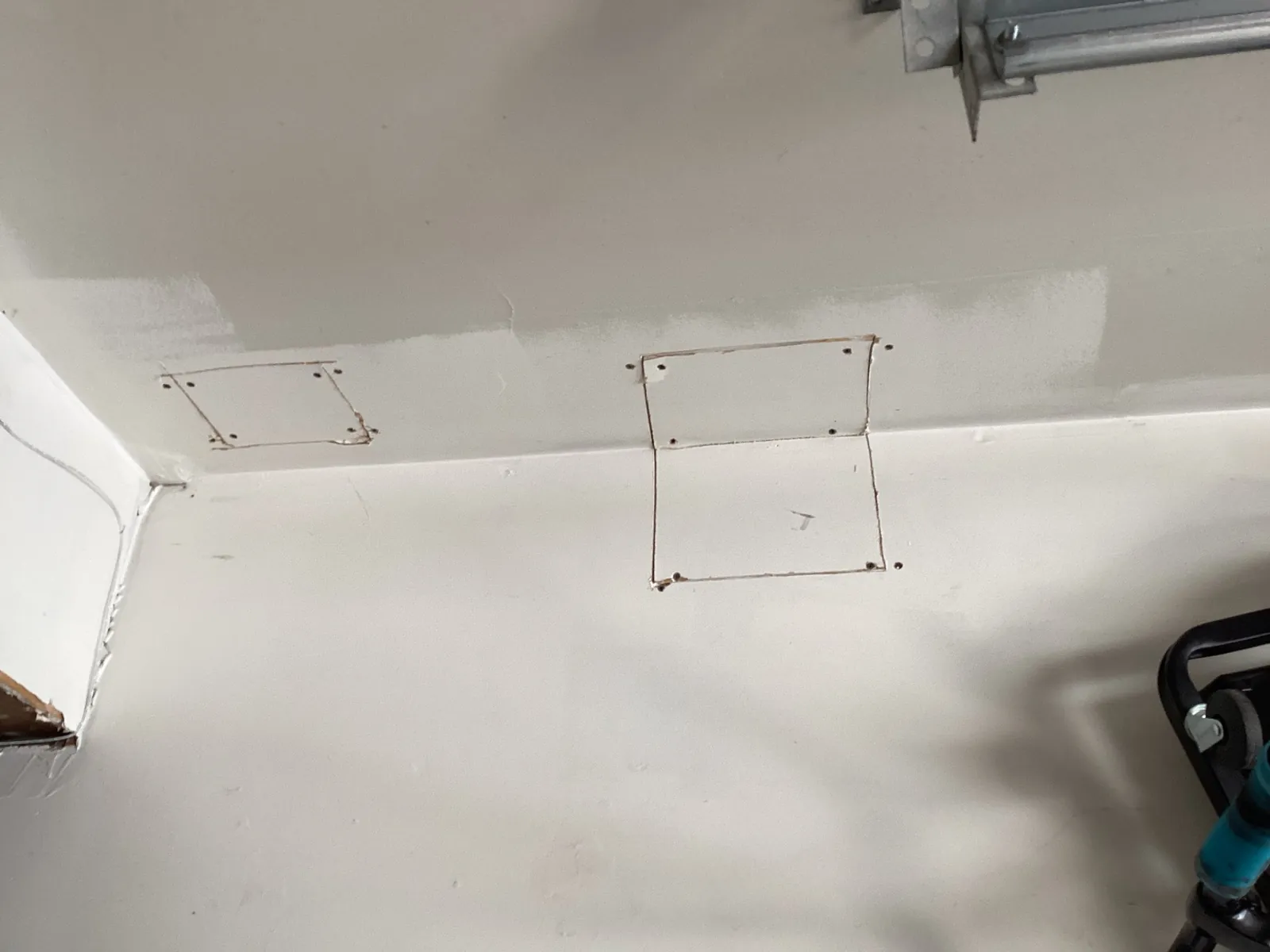 Garage wall and ceiling with square drywall patches screwed in place but never taped or finished, with visible gaps at the edges and one patch folding across the wall-to-ceiling corner