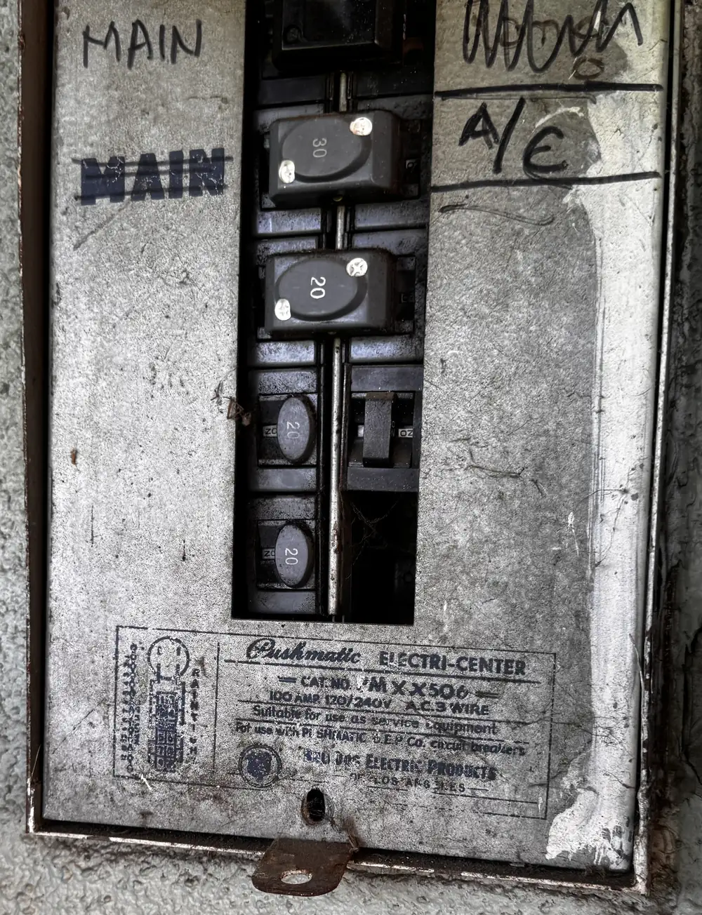 Weathered gray electrical panel with a column of black push-button circuit breakers, hand-written labels reading MAIN and A/C, and a stenciled Pushmatic Electri-Center label